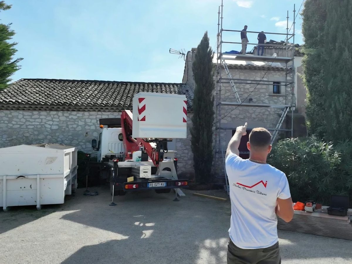 Nicolas désigne la toiture d'un mas en pierre où deux couvreurs interviennent depuis un échafaudage, camion nacelle au premier plan